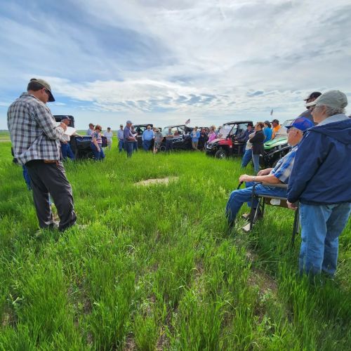 A group of people gathered outdoors in a grassy area, listening to a speaker near parked vehicles under a cloudy sky.