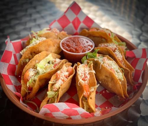 A platter of crispy tacos filled with lettuce, tomatoes, and cheese, served with a side of salsa in a red-checkered basket.