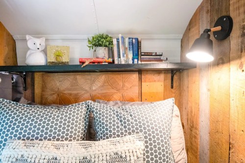Cozy bedroom corner with a wooden wall, decorative pillows, a shelf of books, and a lamp.