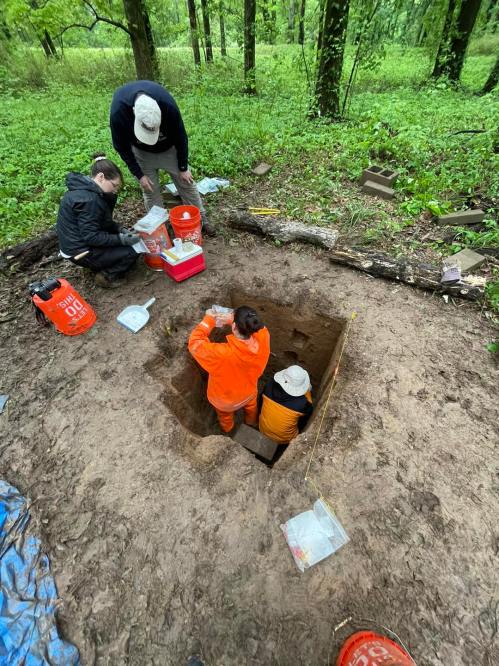 A group of people excavating a square pit in a forested area, surrounded by tools and equipment.