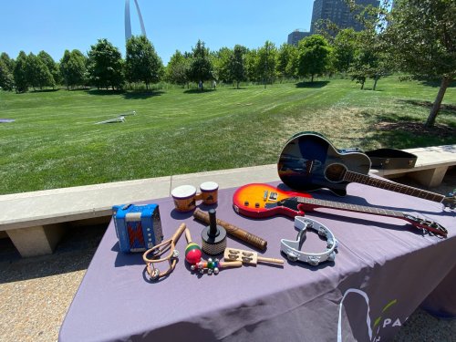A table displays various musical instruments, including guitars, drums, and percussion, set in a sunny park.