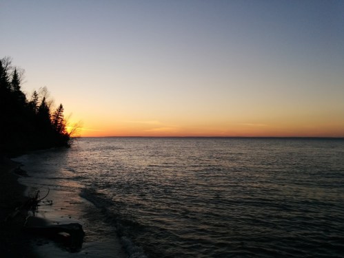 A serene sunset over a calm lake, with silhouettes of trees along the shoreline and a gradient sky.