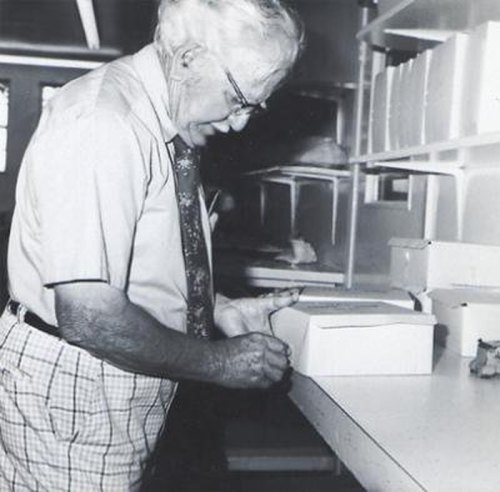 An elderly man in glasses and a tie writes on a box while standing at a table in a bright room.