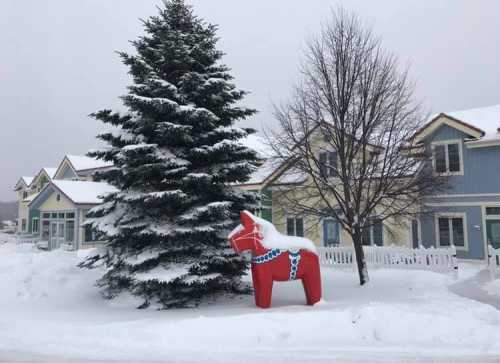 A snowy scene featuring a large red horse statue beside a snow-covered tree and colorful houses in the background.