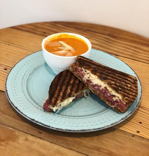 A grilled sandwich with sauerkraut and corned beef, served with a bowl of tomato soup and pickled onions on a blue plate.