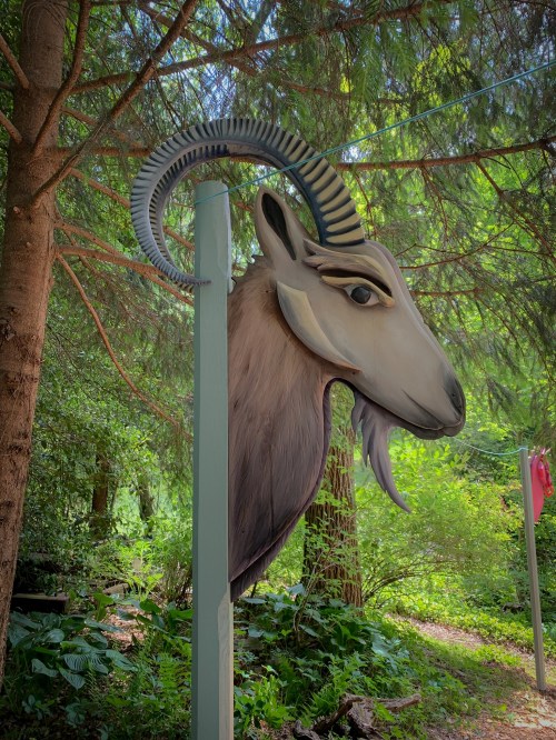 A large, stylized goat head sculpture mounted on a post, surrounded by lush green trees and foliage.