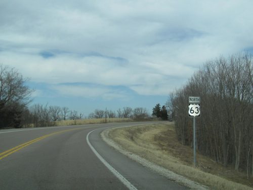 A winding road with a "North 63" sign, surrounded by bare trees and a cloudy sky.