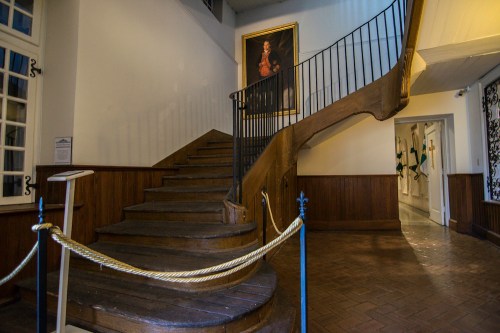 A wooden staircase with a railing, leading to an upper level, beside a wall with a large portrait and a doorway.