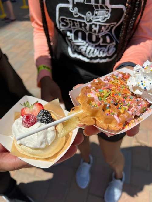 Two hands hold desserts: a mini waffle topped with sprinkles and whipped cream, and a fruit tart with berries and cream.