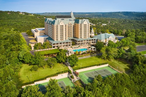 Aerial view of a large hotel surrounded by greenery, with tennis courts and a pool in the foreground.