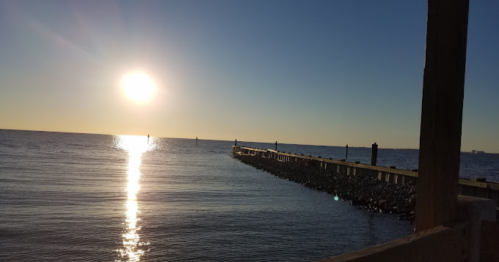 A serene sunset over calm waters, with a pier extending into the horizon and gentle reflections on the surface.