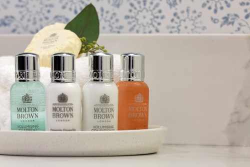 Four Molton Brown travel-sized toiletries in a bathroom setting, featuring shampoo, conditioner, and body wash.