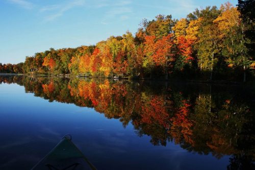 A serene lake reflects vibrant autumn trees in shades of orange, red, and yellow under a clear blue sky.