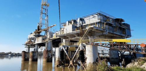 An industrial oil rig stands on stilts over water, with a clear blue sky in the background.