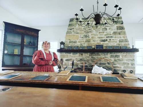 A woman in a historical dress stands by a wooden table with chalkboards, in a room with a stone fireplace and shelves.