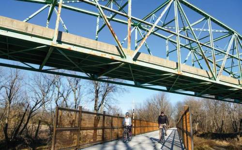 Two cyclists ride on a path beneath a green metal bridge, surrounded by bare trees and clear blue skies.