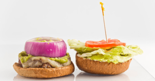 A close-up of two burger halves: one with a beef patty, onion, guacamole, and cheese; the other with lettuce and tomato.