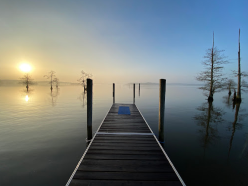 A serene dock extends into a calm lake at sunrise, surrounded by mist and silhouetted trees.