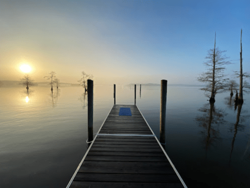 A serene dock extends into a calm lake at sunrise, surrounded by mist and silhouetted trees.