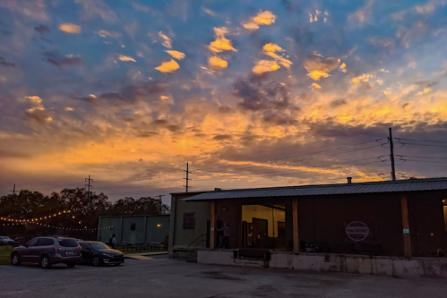 A vibrant sunset with orange and purple hues over a building, with cars parked and string lights in the foreground.