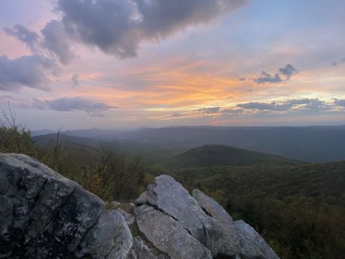A scenic view of rolling hills and a colorful sunset, with rocky foreground and clouds in the sky.