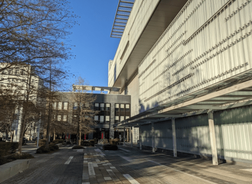Modern building with a glass facade, surrounded by trees and a clear blue sky. Pathway leads to the entrance.