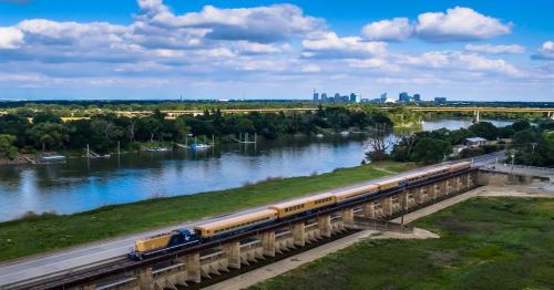 A train travels along a riverbank with a city skyline in the background under a blue sky with fluffy clouds.