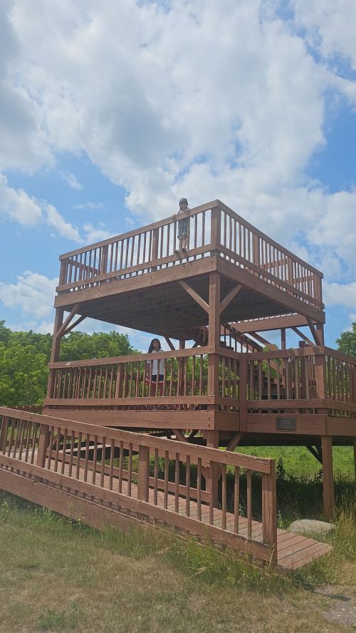 A wooden observation deck with two people, one standing on top and the other below, against a blue sky with clouds.