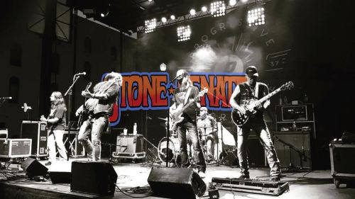 A black and white photo of a band performing on stage with a colorful "STONE TWENTY" graphic overlay.