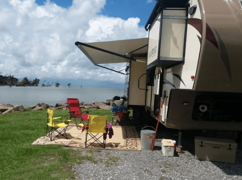 A parked RV by a lake, with two colorful chairs and a rug set up outside under an awning.