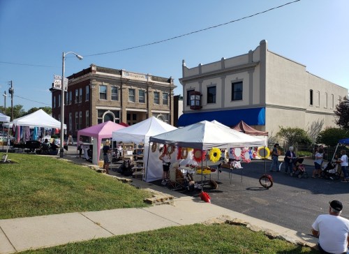 A vibrant outdoor market with tents, vendors, and visitors, set against historic buildings under a clear blue sky.