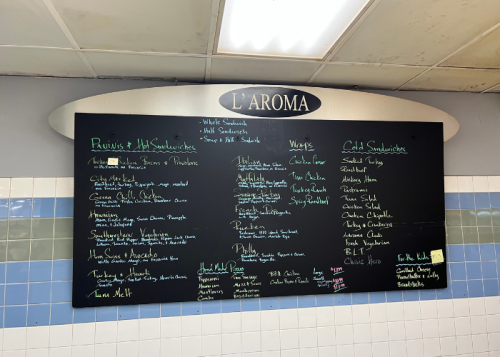Menu board at L'Aroma featuring hot and cold sandwiches, wraps, and soups, with items listed in colorful chalk.