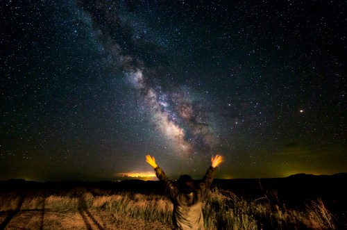 A person stands with arms raised, gazing at a starry night sky filled with the Milky Way and distant lights.