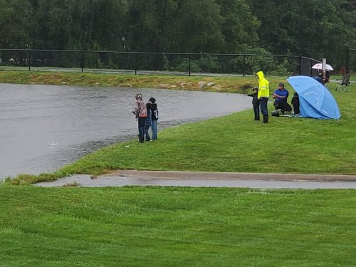 Two people fish by a rain-soaked pond while others sit under an umbrella nearby on a grassy bank.