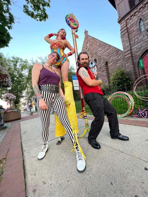 Three performers pose on a sidewalk: one on stilts, two in colorful outfits, surrounded by hula hoops and festive decor.