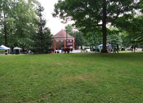 A park scene featuring a bandstand, green grass, trees, and people enjoying a community event.