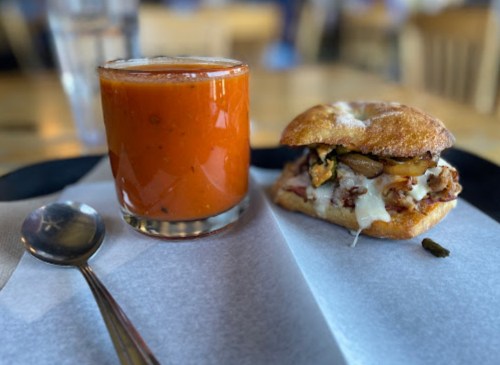 A sandwich with chicken and vegetables next to a glass of tomato soup on a tray with a spoon.