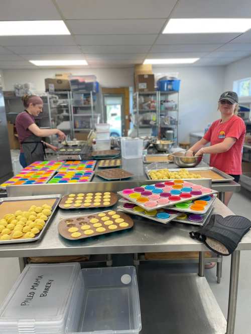 Two bakers work in a bright kitchen, surrounded by trays of colorful desserts and baking supplies.