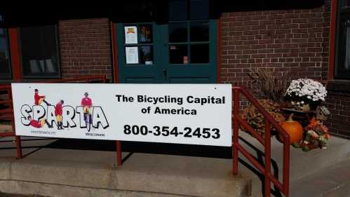 Sign outside a building reading "Sparta, The Bicycling Capital of America" with flowers and pumpkins nearby.