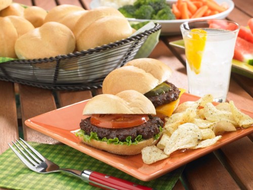 A plate with two mini burgers, lettuce, tomato, and pickles, served with chips and a glass of lemonade.