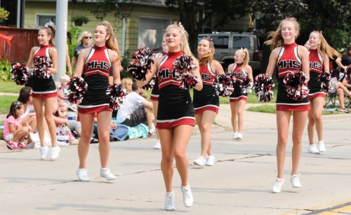 A group of cheerleaders in black and red uniforms perform with pom-poms during a parade, with spectators in the background.