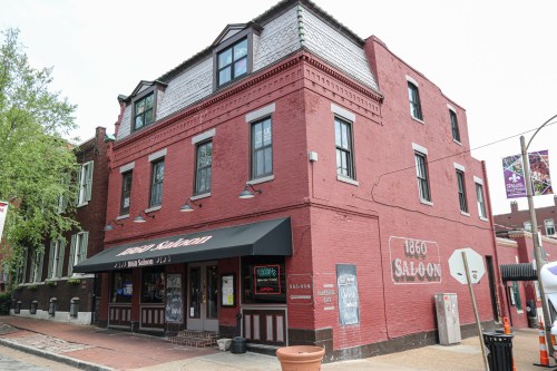 Exterior of the 1860 Saloon, a red brick building with a black awning and open sign, surrounded by trees and street elements.