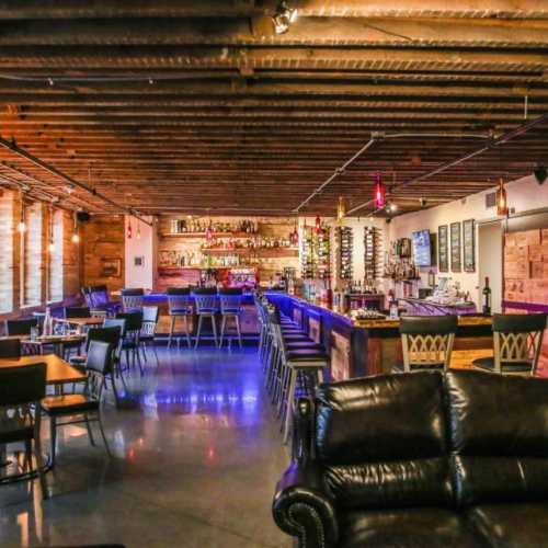 A stylish bar interior with wooden beams, a long bar, seating areas, and shelves filled with bottles.