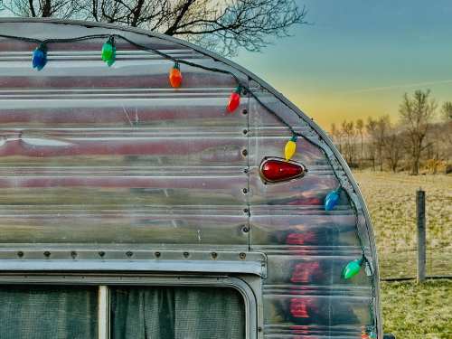 A vintage trailer adorned with colorful holiday lights, set against a serene landscape at sunset.