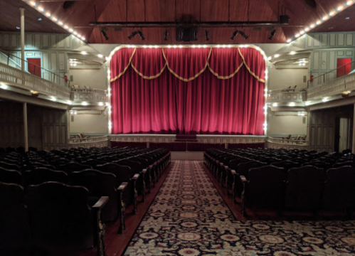 A grand theater interior with red curtains, ornate lighting, and empty rows of seats facing the stage.