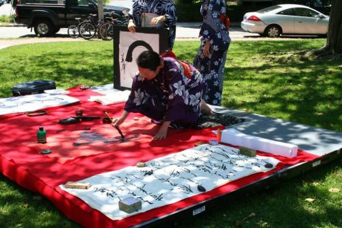 A woman in a kimono paints calligraphy on a red mat, surrounded by traditional art supplies and two others in the background.