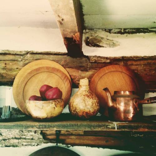 Wooden plates and a gourd sit on a rustic shelf, alongside a copper teapot and a bowl of red apples.