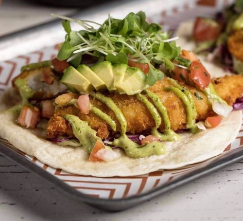 Two fish tacos topped with avocado, pico de gallo, microgreens, and drizzled with green sauce on a tray.