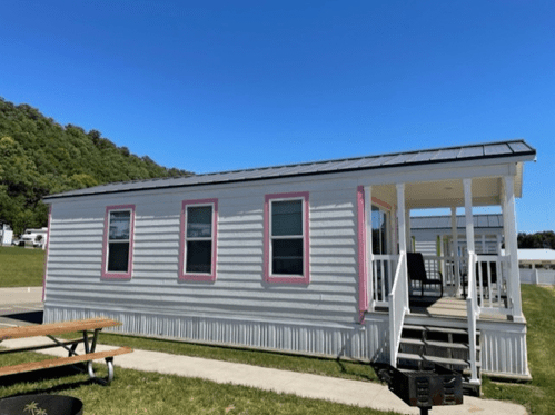 A modern mobile home with a porch, set against a clear blue sky and green hills in the background.
