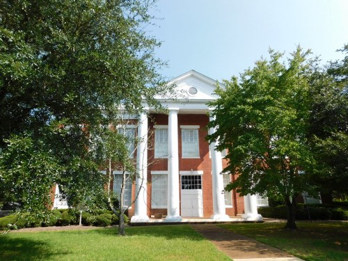 A brick building with white columns, surrounded by green trees and a manicured lawn under a clear blue sky.